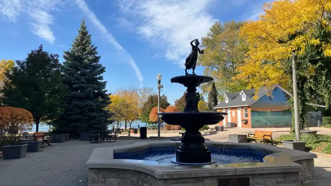 Itim na fountain sa Amherstburg waterfront park na may mga puno ng taglagas at ang Detroit River sa likod nito